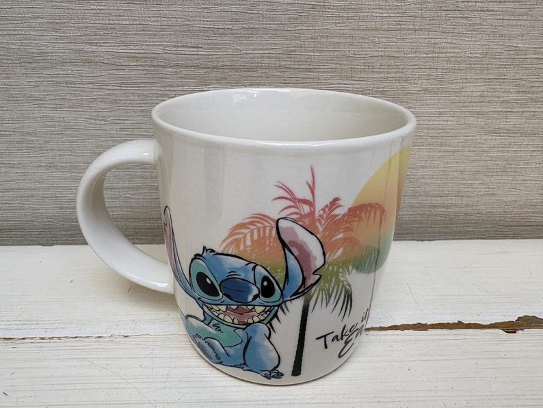 May include: White ceramic mug featuring a cartoon image of Stitch from Lilo & Stitch. The mug has a curved handle and a colorful design of Stitch, palm trees, and a sunset. The text "Take it Easy" is also visible.