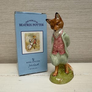 May include: A porcelain figurine of a fox dressed in a pink waistcoat and light green jacket, standing on a green base. Beside it is a light blue box labelled The World of Beatrix Potter and Royal Doulton, featuring an illustration of a fox and a duck.