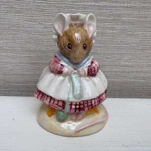 Könnte beinhalten: Keramikfigur einer Maus mit Häubchen und Kleid. Die Maus hat braunes Fell, rosa Ohren und trägt ein weißes Kleid mit rosa und roter Bordüre. Sie hält einen grünen Gegenstand und steht auf einem beigen Sockel.