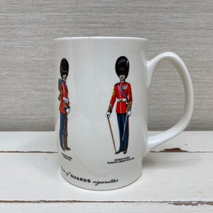 May include: White ceramic mug with a red, white, and blue illustration of two British guards in uniform. The text "Guards cigarettes" is printed on the bottom of the mug.