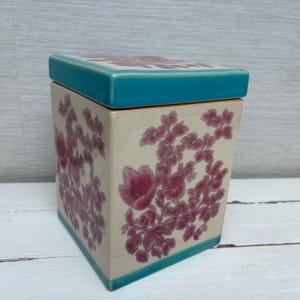 May include: Square ceramic box with a teal lid and a floral design in shades of pink and white. The box has a floral pattern on all sides.