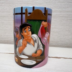 May include: A cylindrical ceramic mug featuring a colorful illustration of a scene from a Disney animated film. The design includes a male character in a white shirt, plates, and other characters in the background. The mug has a light blue top and a pink base.