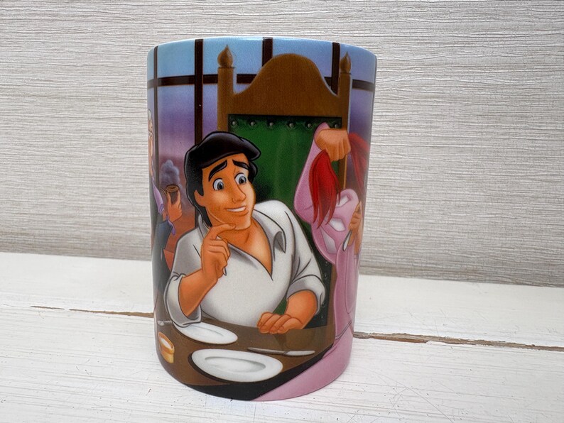 May include: A cylindrical ceramic mug featuring a colorful illustration of a scene from a Disney animated film. The design includes a male character in a white shirt, plates, and other characters in the background. The mug has a light blue top and a pink base.