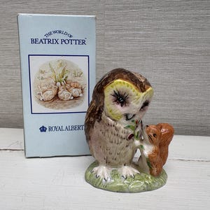 May include: A ceramic figurine of a brown owl and a small orange squirrel standing on a green base, positioned next to a light blue rectangular box labelled The World of Beatrix Potter and Royal Albert with a circular illustration of rabbits.