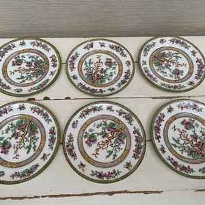 Könnte beinhalten: Sechs weiße Porzellanplatten mit grünem und goldenem Rand und einem floralen Design in der Mitte. Das florale Design zeigt einen rosa und grünen Ast mit rosa Blüten.