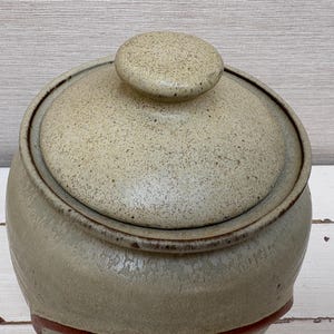 May include: A ceramic jar with a lid. The jar is a light beige color with a speckled texture. The lid has a small, round knob on top. The jar has a dark brown rim and a red band near the base. The background is a light wood.
