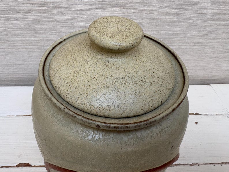 May include: A ceramic jar with a lid. The jar is a light beige color with a speckled texture. The lid has a small, round knob on top. The jar has a dark brown rim and a red band near the base. The background is a light wood.