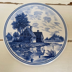 May include: Blue and white ceramic tile featuring a Dutch countryside scene. The tile depicts a small house with a blue roof, a tree, and a river. The tile is surrounded by a white border.