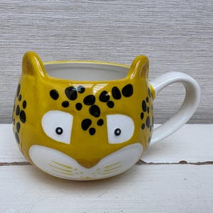 Peut inclure: Une tasse en céramique jaune avec un motif léopard noir et blanc. La tasse a une anse blanche et un visage de guépard de dessin animé mignon avec des yeux noirs et un nez blanc.