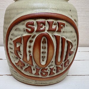 May include: A ceramic jar with a beige base and a brown, oval-shaped design. The design features the words "SELF FLOUR RAISING" in a retro font. The jar has a textured surface and a rounded shape, suitable for kitchen storage.