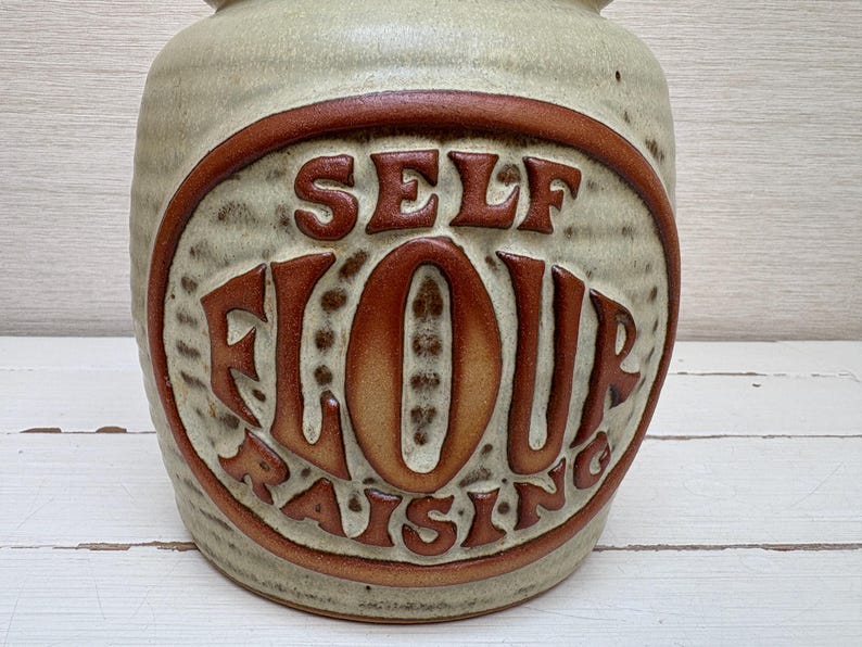 May include: A ceramic jar with a beige base and a brown, oval-shaped design. The design features the words "SELF FLOUR RAISING" in a retro font. The jar has a textured surface and a rounded shape, suitable for kitchen storage.