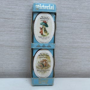 Puede incluir: Una caja azul y blanca con la palabra "Pictorial" impresa en la parte delantera. La caja contiene dos azulejos de cerámica ovalados con ilustraciones de personajes de Beatrix Potter. El azulejo superior presenta el texto "The Tale of Benjamin Bunny" y una ilustración de Benjamin Bunny. El azulejo inferior presenta el texto "The Tale of Mr. Jeremy Fisher" y una ilustración de Mr. Jeremy Fisher.