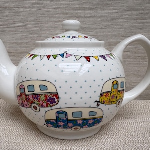 以下が含まれることがあります： 蓋と取っ手が付いた白いセラミックティーポット。 ティーポットは、3つのカラフルなキャンピングカーのイラストと水玉模様の背景で装飾されています。 カラフルなペナントフラッグのストリングが上部を飾っています。