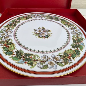 May include: A white ceramic plate with a gold rim features a festive holiday design. The plate is decorated with a wreath of green holly leaves, red berries, and white flowers. The centre of the plate has a floral design with white flowers and green leaves.