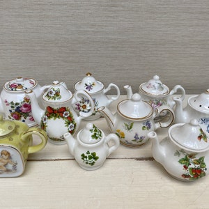 Könnte beinhalten: Eine Sammlung von zehn Miniatur-Porzellan-Teekannen mit floralen und fantasievollen Designs. Die Teekannen sind in verschiedenen Farben, darunter Weiß, Grün und Gelb, und weisen komplizierte Details auf.