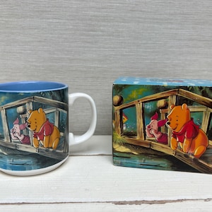 Könnte beinhalten: Eine Keramik-Tasse mit blauem Innenraum und weißem Henkel, mit einer farbenfrohen Illustration von Winnie Puuh und Ferkel auf einer Brücke. Eine passende Schachtel befindet sich neben der Tasse.
