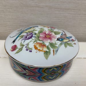 Può includere: Scatola portagioie rotonda in ceramica bianca con coperchio decorato con fiori e uccelli. Il coperchio presenta fiori rosa, arancioni e viola e uccelli colorati. La base ha un motivo a spirale colorato. Con bordo dorato.