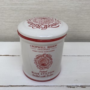 May include: White ceramic jar with a red lid and red lettering. The jar is labeled "Cropwell Bishop Creamery Finest Hand Made Cheese" and "Potted Blue Stilton Sealed with Butter".