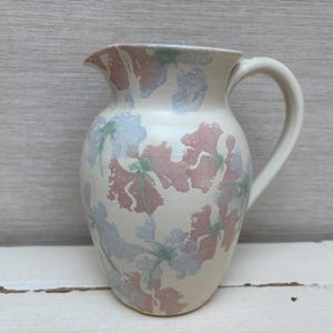 Könnte beinhalten: Eine weiße Keramikkanne mit einem Blumendesign in Rosa-, Blau- und Grüntönen. Die Kanne hat einen Henkel und einen runden Korpus.