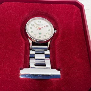 Könnte beinhalten: Eine silberfarbene Krankenschwester-Taschenuhr mit weißem Zifferblatt, schwarzen Ziffern und rotem Sekundenzeiger, präsentiert in einer roten Samtbox. Die Uhr hat ein klassisches Design mit Kettenbefestigung.