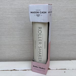 Può includere: Un Mason Cash Roller Shaker nella sua confezione originale. Lo shaker bianco sporco è in una scatola rosa e bianca. Il testo "ROLLER SHAKER" è stampato verticalmente sullo shaker. La scatola recita "MASON CASH ENGLAND SINCE 1800".