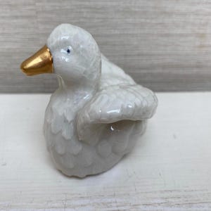 Könnte beinhalten: Eine weiße Keramik-Entenfigur mit goldenem Schnabel. Die Ente hat eine strukturierte Oberfläche und blaue Augen. Die Figur ist ein dekorativer Artikel.