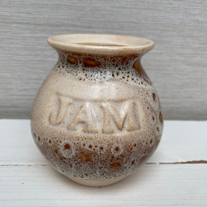 Puede incluir: Un pequeño tarro de cerámica blanco con motas marrones y la palabra "JAM" en relieve en la parte delantera.
