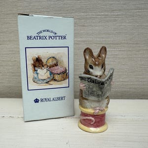 May include: A ceramic figurine of a brown mouse reading a newspaper titled The Tailor Gloucester, perched on a pink thread spool. Beside it stands a light blue cardboard box labeled The World of Beatrix Potter and Royal Albert with an illustration.
