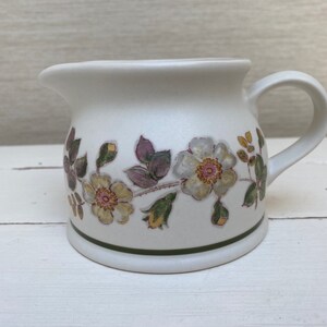 Könnte beinhalten: Eine weiße Keramik-Sahnekännchen mit floralem Muster. Das Sahnekännchen hat einen Griff und einen Ausguss zum Ausgießen. Das florale Muster umfasst weiße, gelbe und violette Blumen mit grünen Blättern. Ein grünes Band umrandet den Boden.
