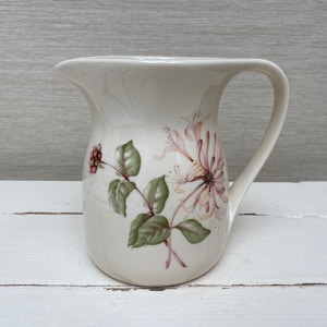 Könnte beinhalten: Eine weiße Keramikkanne mit einem Blumendesign mit rosa und gelben Geißblattblumen und grünen Blättern. Die Kanne hat einen Henkel und einen runden Körper.