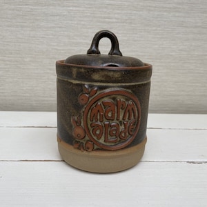 Könnte beinhalten: Eine braune Keramikdose mit Deckel und braunem Henkel. Die Dose hat ein braunes und orangefarbenes Design mit den Worten "farm made" auf der Vorderseite.