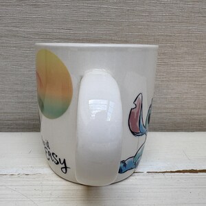 May include: White ceramic mug featuring a colorful illustration of Stitch from Lilo & Stitch. The mug has a curved handle and the words "make it easy" in black script. The design includes a sunset-like graphic.