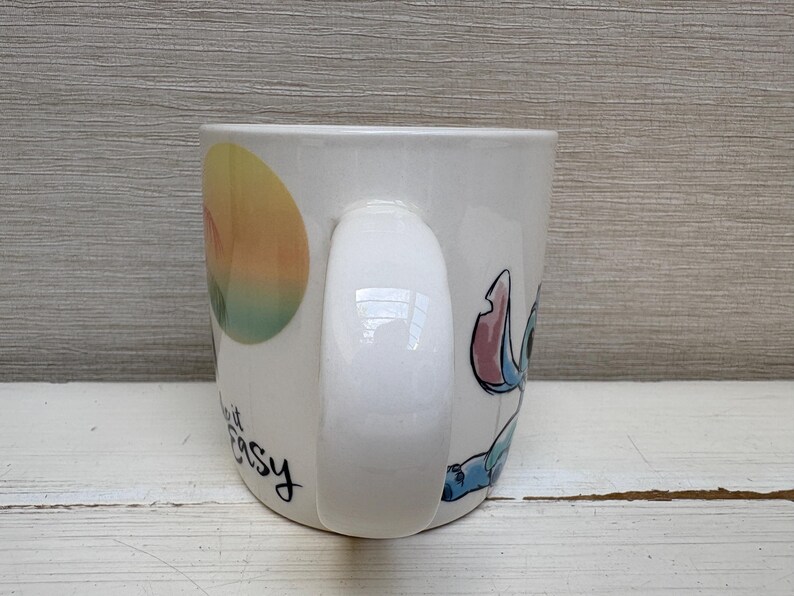 May include: White ceramic mug featuring a colorful illustration of Stitch from Lilo & Stitch. The mug has a curved handle and the words "make it easy" in black script. The design includes a sunset-like graphic.