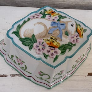 Könnte beinhalten: Weiße Keramikschale mit Lamm- und Blumendesign. Das Lamm hat eine blaue Schleife und ist von rosa Blumen und Schmetterlingen umgeben. Die Schale hat einen gewellten Rand mit türkisfarbenem Rand und floralen Akzenten.