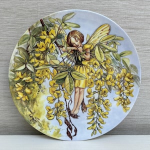 Può includere: Piatto decorativo con una fata che riposa tra fiori gialli e foglie verdi. La fata ha i capelli castani, ali gialle e un vestito giallo. Il piatto ha uno sfondo azzurro chiaro e un bordo floreale.