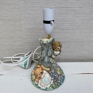 Peut inclure: Une lampe de table en céramique en forme de tronc d'arbre avec une petite chouette et un lapin en figurine. La lampe a un abat-jour blanc et un cordon blanc.
