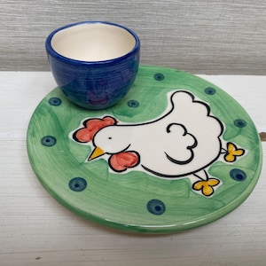 Könnte beinhalten: Ein Keramik-Eierbecher- und Teller-Set. Der blaue Eierbecher sitzt auf einem grünen Teller, der mit einer weißen Hühnerillustration verziert ist. Der Teller hat einen grünen Rand mit blauen Punkten. Das Huhn hat einen roten Kamm und einen gelben Schnabel.