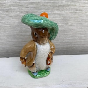 Puede incluir: Figura de cerámica de un conejo con un sombrero verde con un acento naranja. El conejo es marrón y blanco, sosteniendo una pequeña bolsa. La figura está sobre una base verde.