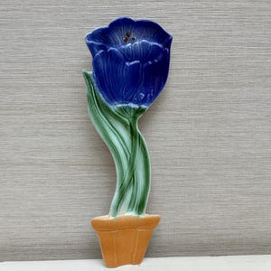 Könnte beinhalten: Eine keramische Tulpen-Dekoration. Die Blüte ist tiefblau, der Stiel grün und weiß und der Topf orange. Das Stück ist zum Aufstellen konzipiert und dient wahrscheinlich zur Dekoration.