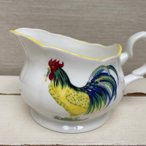 May include: White ceramic creamer with a yellow, blue and green rooster design. The creamer has a scalloped edge and a handle.