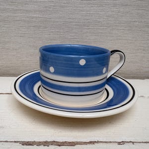 Puede incluir: Un juego de taza y platillo de té a rayas azules y blancas con lunares blancos. La taza y el platillo son de cerámica.