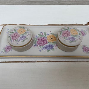 Könnte beinhalten: Drei kleine, runde, weiße Keramik-Schmuckkästchen mit Blumendesigns. Die Kästchen befinden sich auf einem weißen Tablett mit goldener Umrandung.