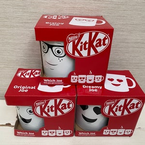 May include: Three red and white cardboard boxes with different designs of white mugs with faces on them. The boxes are stacked on top of each other. The boxes have the Nestle KitKat logo on them. The text on the boxes reads "Original Joe", "Brainy Joe", and "Dreamy Joe".
