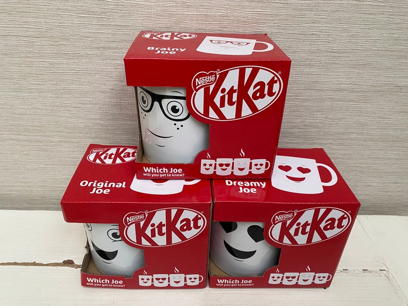 May include: Three red and white cardboard boxes with different designs of white mugs with faces on them. The boxes are stacked on top of each other. The boxes have the Nestle KitKat logo on them. The text on the boxes reads "Original Joe", "Brainy Joe", and "Dreamy Joe".