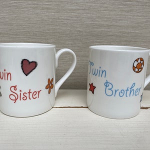 May include: Two white ceramic mugs with handles. One mug has the words "Twin Sister" in red script, with a heart and flower illustrations. The other mug says "Twin Brother" in blue script, with a star and a ball illustration.