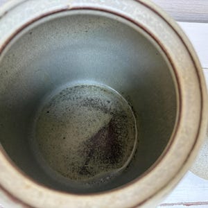 May include: A ceramic jar with a lid, featuring a speckled beige exterior and a dark brown interior. The jar has a round shape with a lid on the side. The interior has a dark brown and gray speckled pattern.