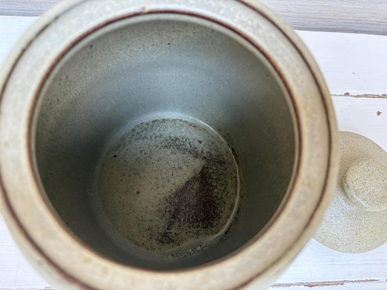 May include: A ceramic jar with a lid, featuring a speckled beige exterior and a dark brown interior. The jar has a round shape with a lid on the side. The interior has a dark brown and gray speckled pattern.