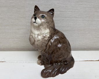 May include: A brown and white ceramic figurine of a cat sitting with its head tilted up. The cat has a fluffy tail and a white patch on its chest.