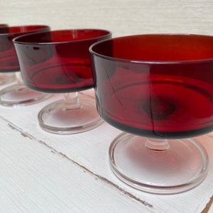 Könnte beinhalten: Satz mit vier roten gläsernen Desserttellern mit klaren Glasstielen und -sockeln. Die Teller sind in einer Reihe auf einer weißen Oberfläche angeordnet.