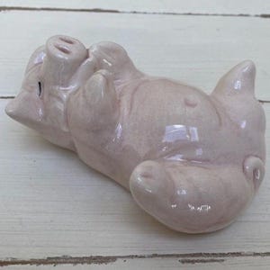 Puede incluir: Figurita de cerdo de cerámica rosa claro, tumbada boca arriba con las patas en el aire. El cerdo tiene detalles, incluyendo un hocico, orejas y una cola enroscada. La figurita está sobre una superficie de madera blanca.